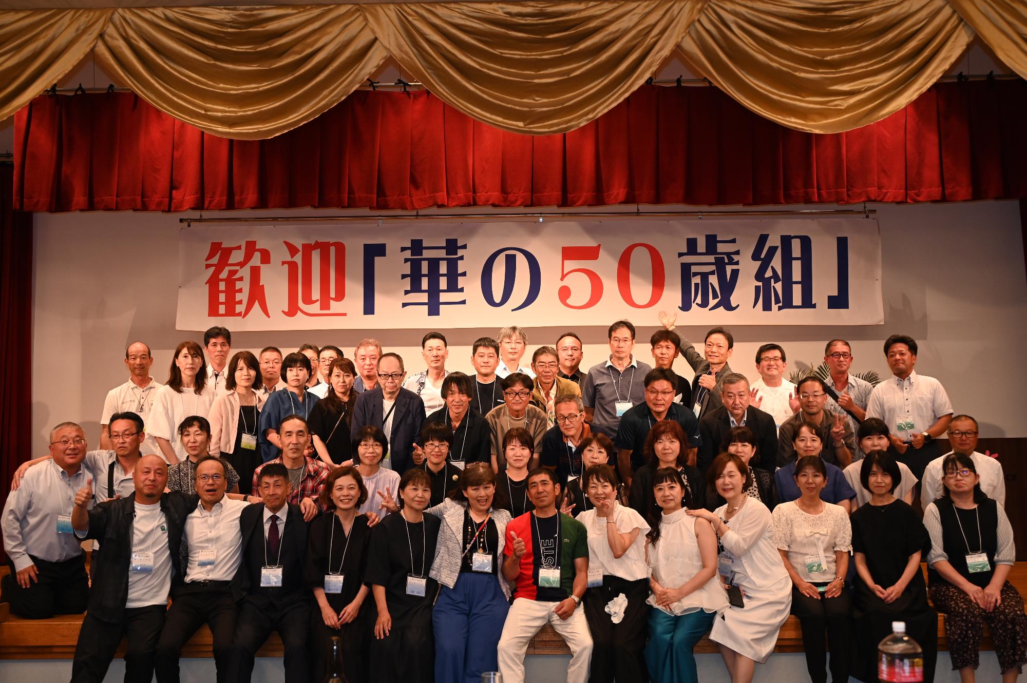 「歓迎 華の50歳組」と書かれた横断幕の前に並ぶ参加者たちの集合写真（阿久根小）
