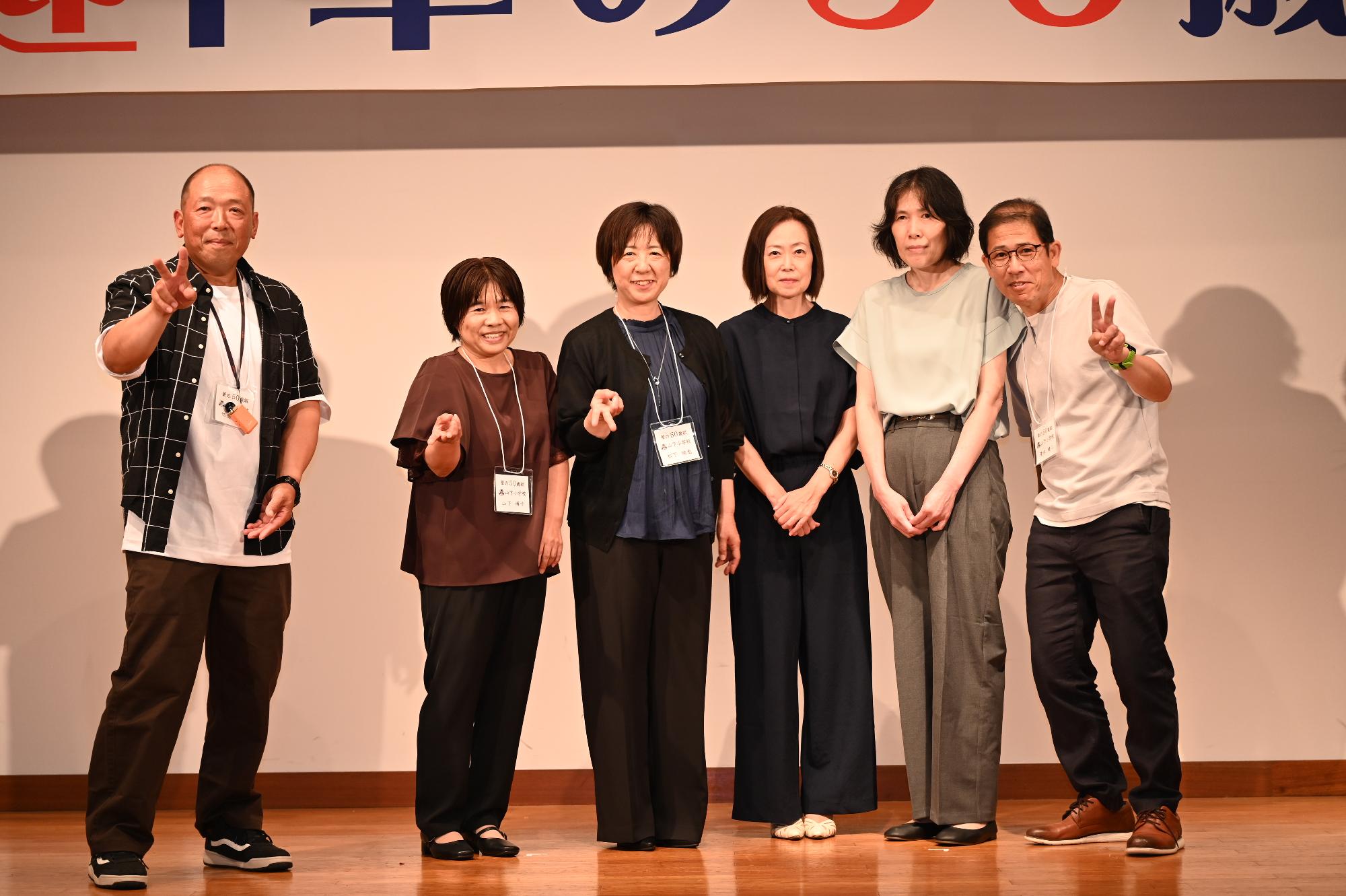 「歓迎 華の50歳組」と書かれた横断幕の前に並ぶ参加者たちの集合写真（山下小）
