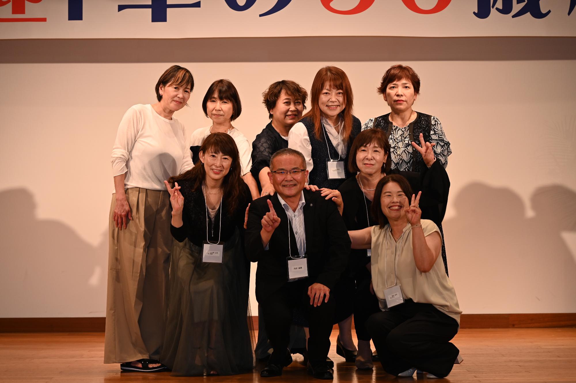 「歓迎 華の50歳組」と書かれた横断幕の前に並ぶ参加者たちの集合写真（西目小）