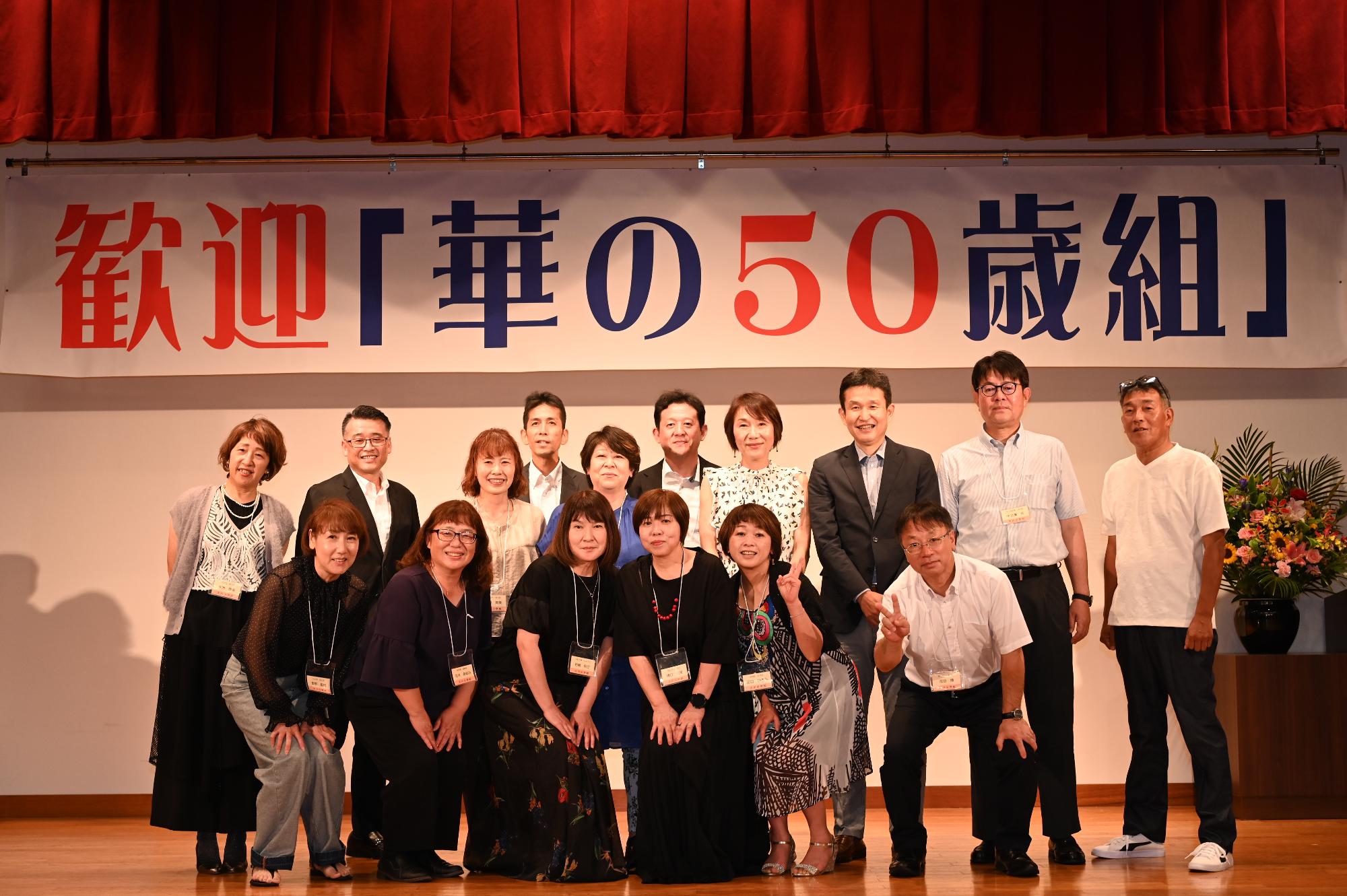 「歓迎 華の50歳組」と書かれた横断幕の前に並ぶ参加者たちの集合写真（大川小）