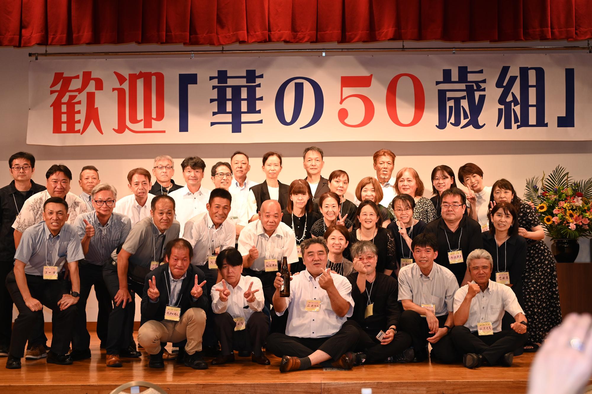 「歓迎 華の50歳組」と書かれた横断幕の前に並ぶ参加者たちの集合写真（脇本小）