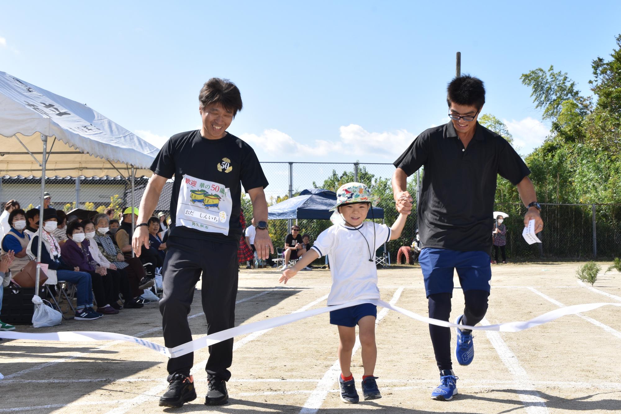 間に子供をはさんで手をつなぎながらゴールをする2人の男性の写真