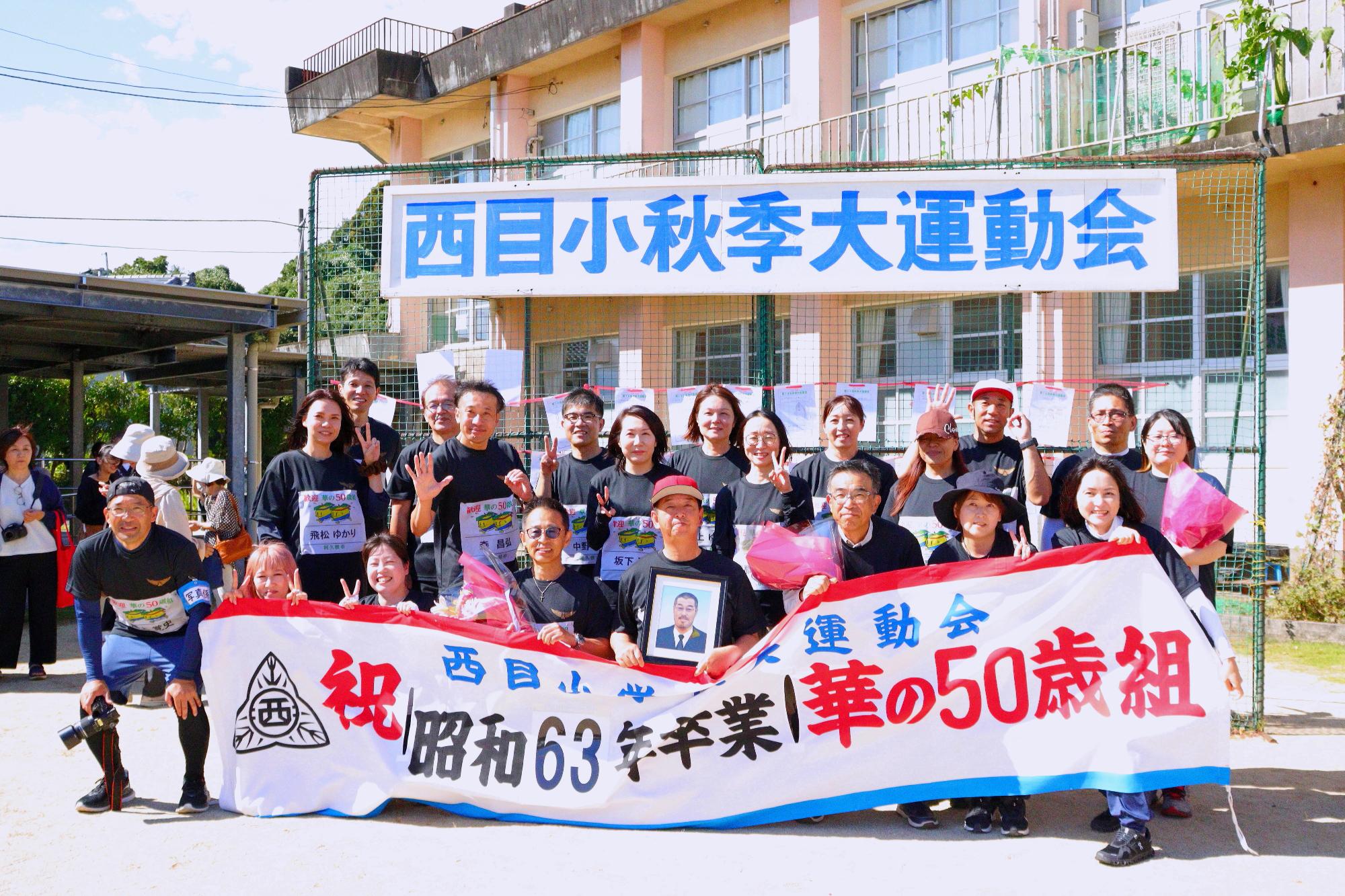 華の50歳組の前に掲げて集合写真を撮る西目小学校華の50歳組の参加者