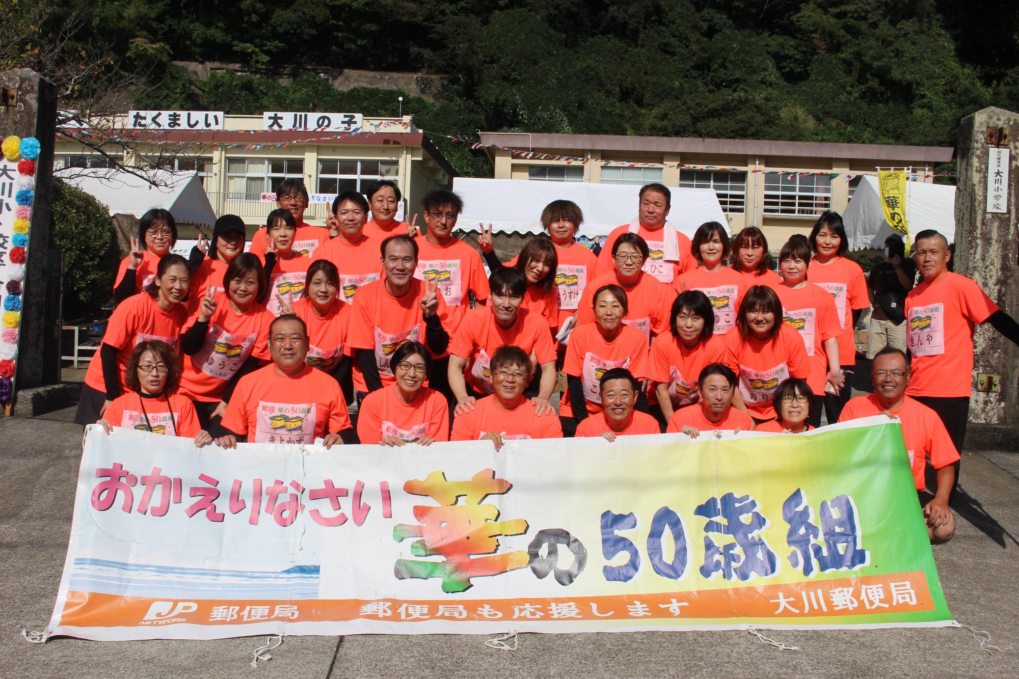 「おかえりなさい華の50歳組」と書かれた横断幕旗を持って集合写真を撮る大川小学校華の50歳組の参加者