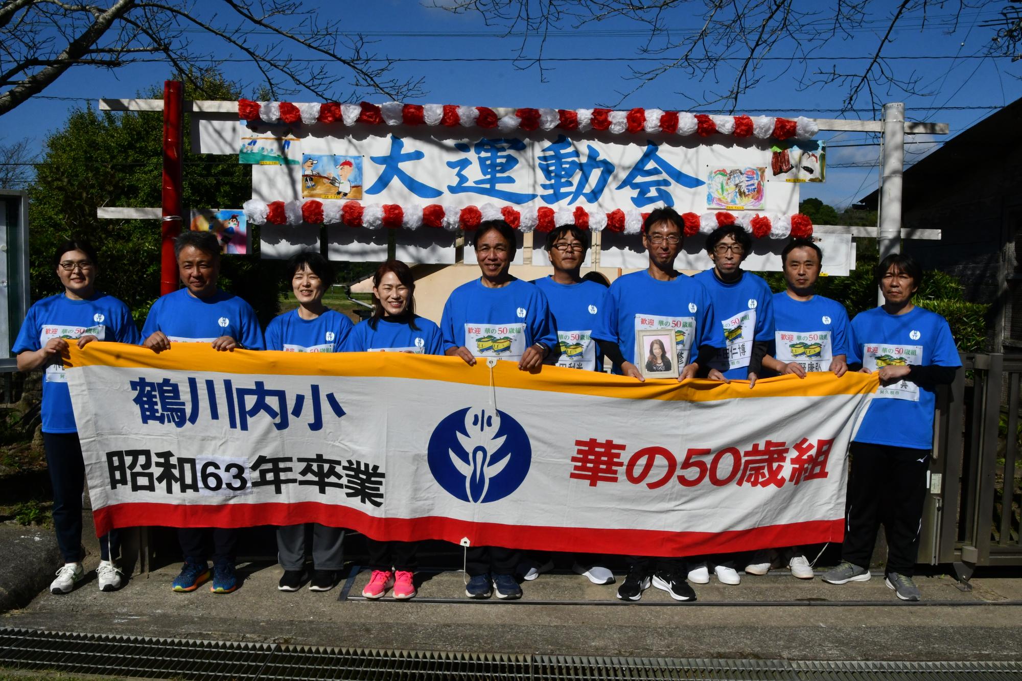 校門前で華の50歳組の横断幕を持ちながら写真を撮る参加者の写真