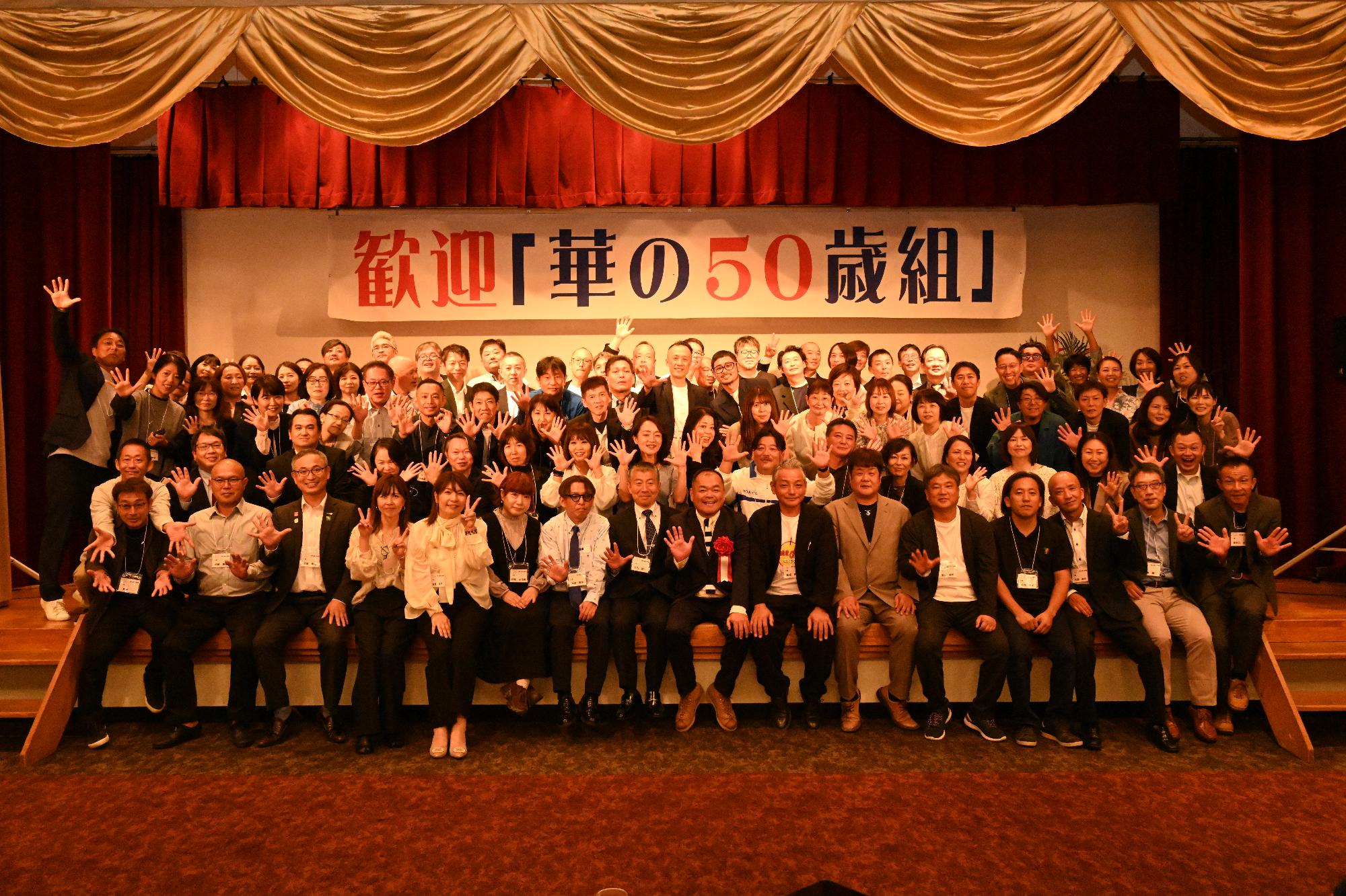 「歓迎 華の50歳組」と書かれた横断幕の前に並ぶ参加者たちの集合写真（阿久根小）