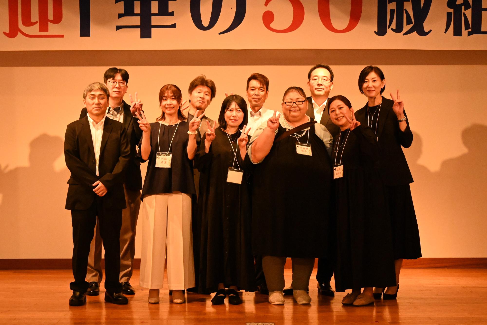 「歓迎 華の50歳組」と書かれた横断幕の前に並ぶ参加者たちの集合写真（山下小）