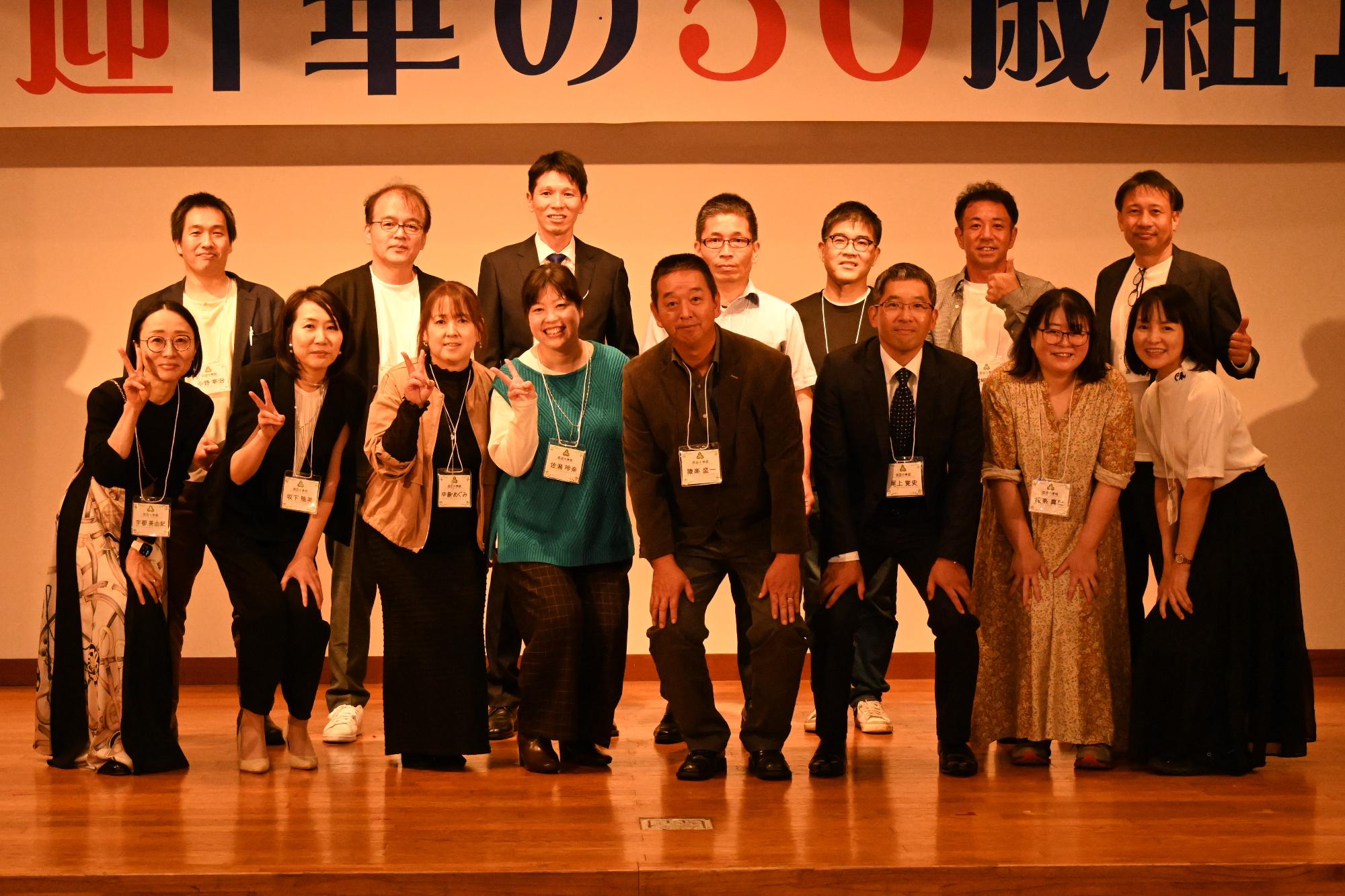 「歓迎 華の50歳組」と書かれた横断幕の前に並ぶ参加者たちの集合写真（西目小）