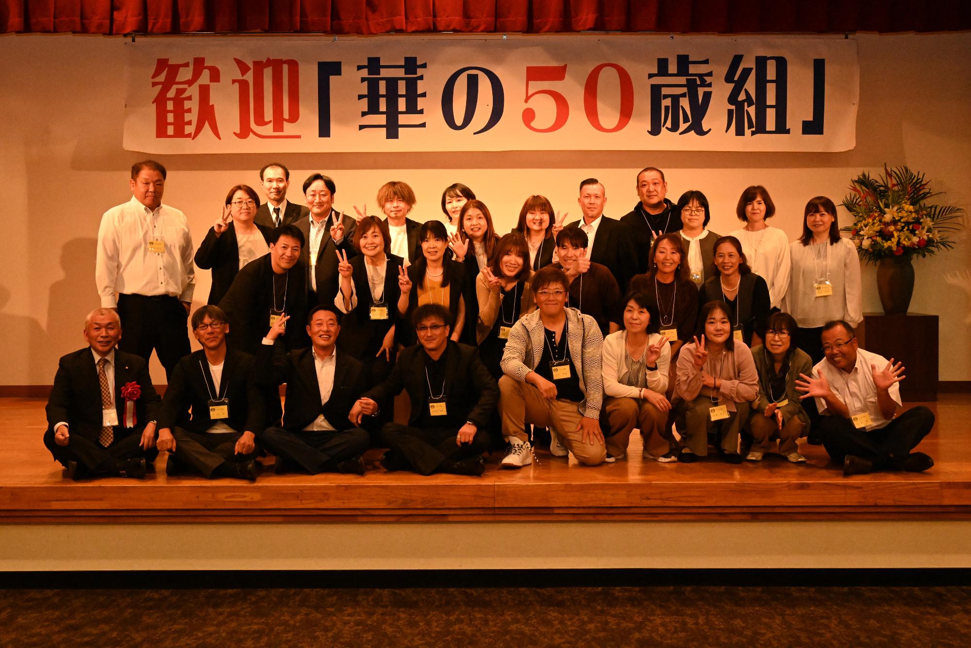 「歓迎 華の50歳組」と書かれた横断幕の前に並ぶ参加者たちの集合写真（大川小）