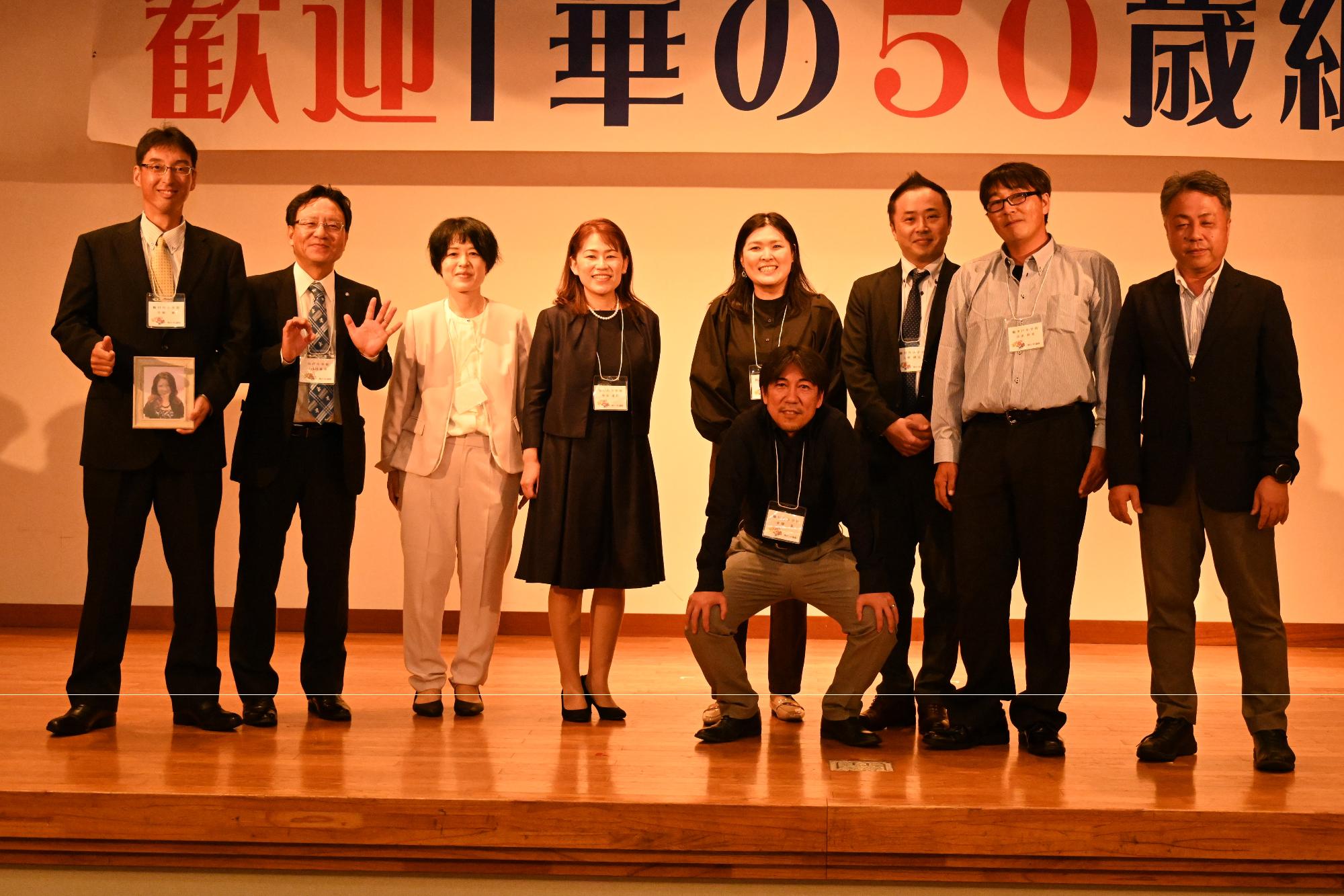 「歓迎 華の50歳組」と書かれた横断幕の前に並ぶ参加者たちの集合写真（鶴川内小）