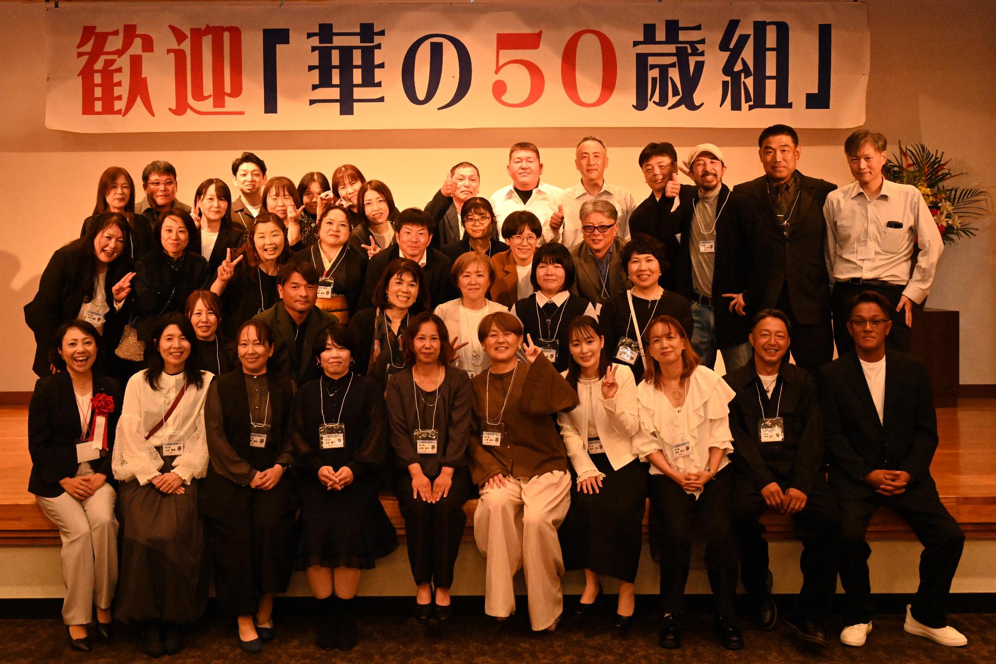 「歓迎 華の50歳組」と書かれた横断幕の前に並ぶ参加者たちの集合写真（脇本小）
