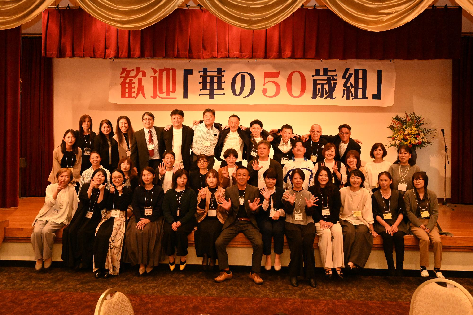 「歓迎 華の50歳組」と書かれた横断幕の前に並ぶ参加者たちの集合写真（阿久根高校出身）