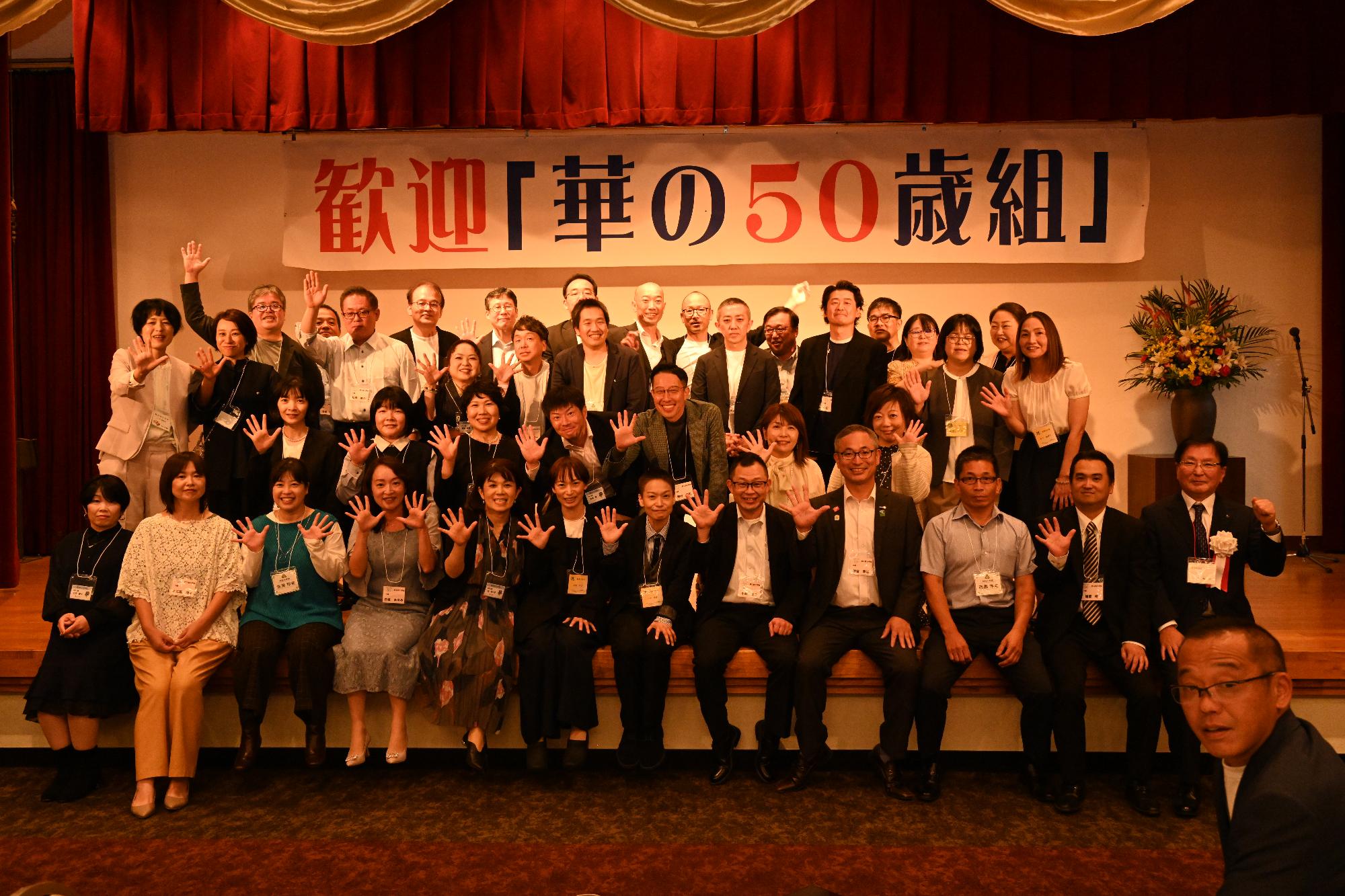 「歓迎 華の50歳組」と書かれた横断幕の前に並ぶ参加者たちの集合写真（出水高校出身）