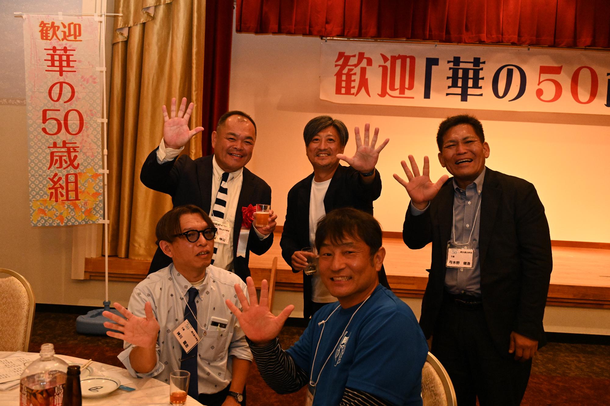 テーブルの前で手で5のポーズをとる阿久根小出身の5人の男性の写真