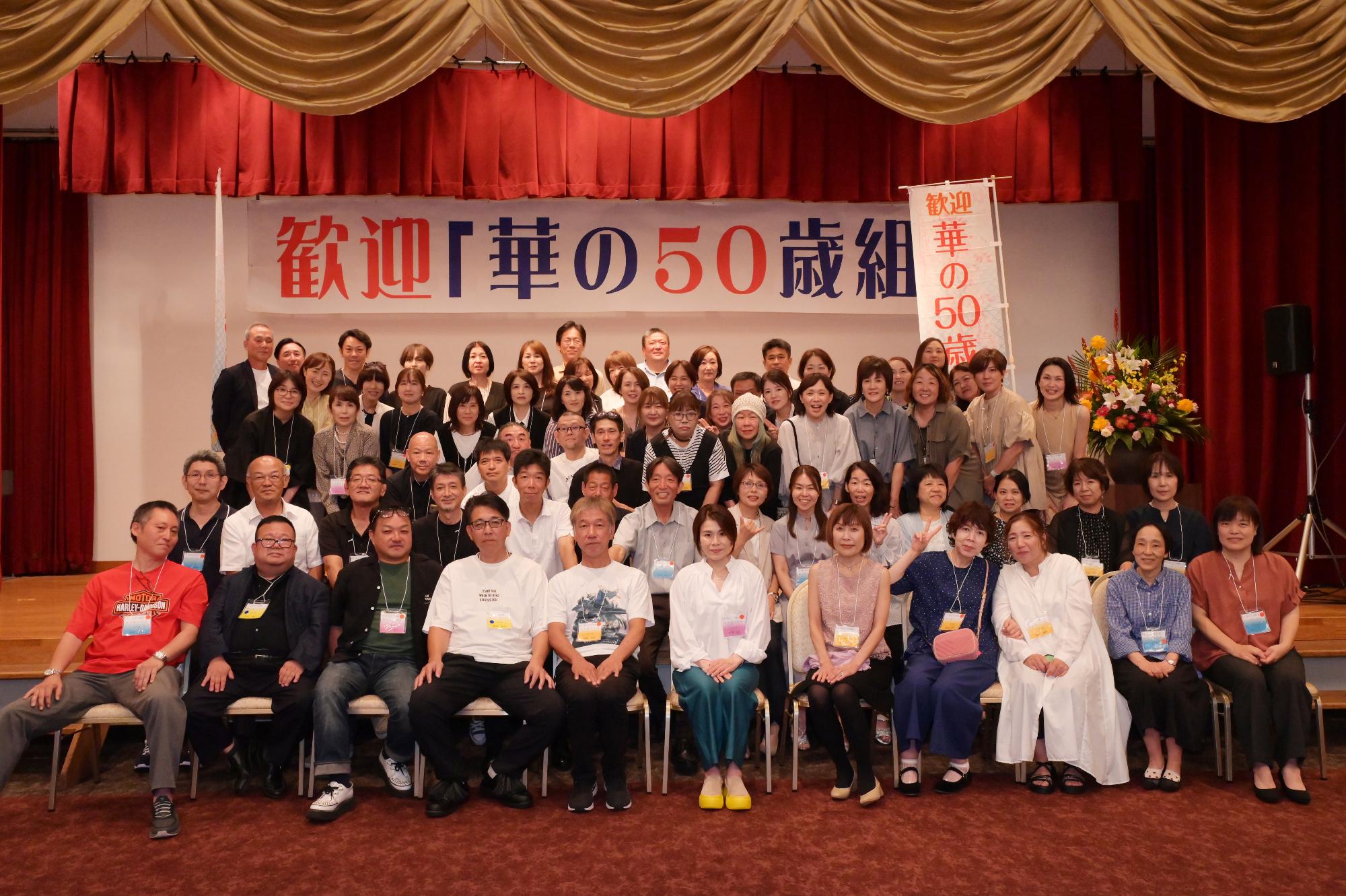 「歓迎 華の50歳組」と書かれた横断幕の前に並ぶ参加者たちの集合写真（阿久根小）