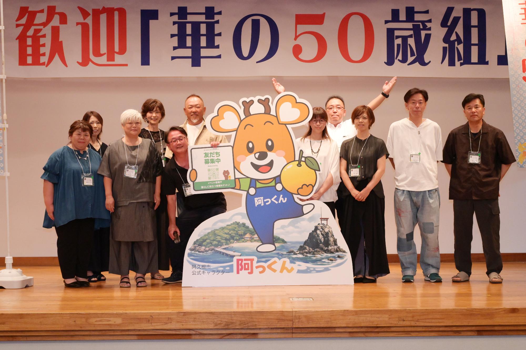 「歓迎 華の50歳組」と書かれた横断幕の前に並ぶ参加者たちの集合写真（山下小）
