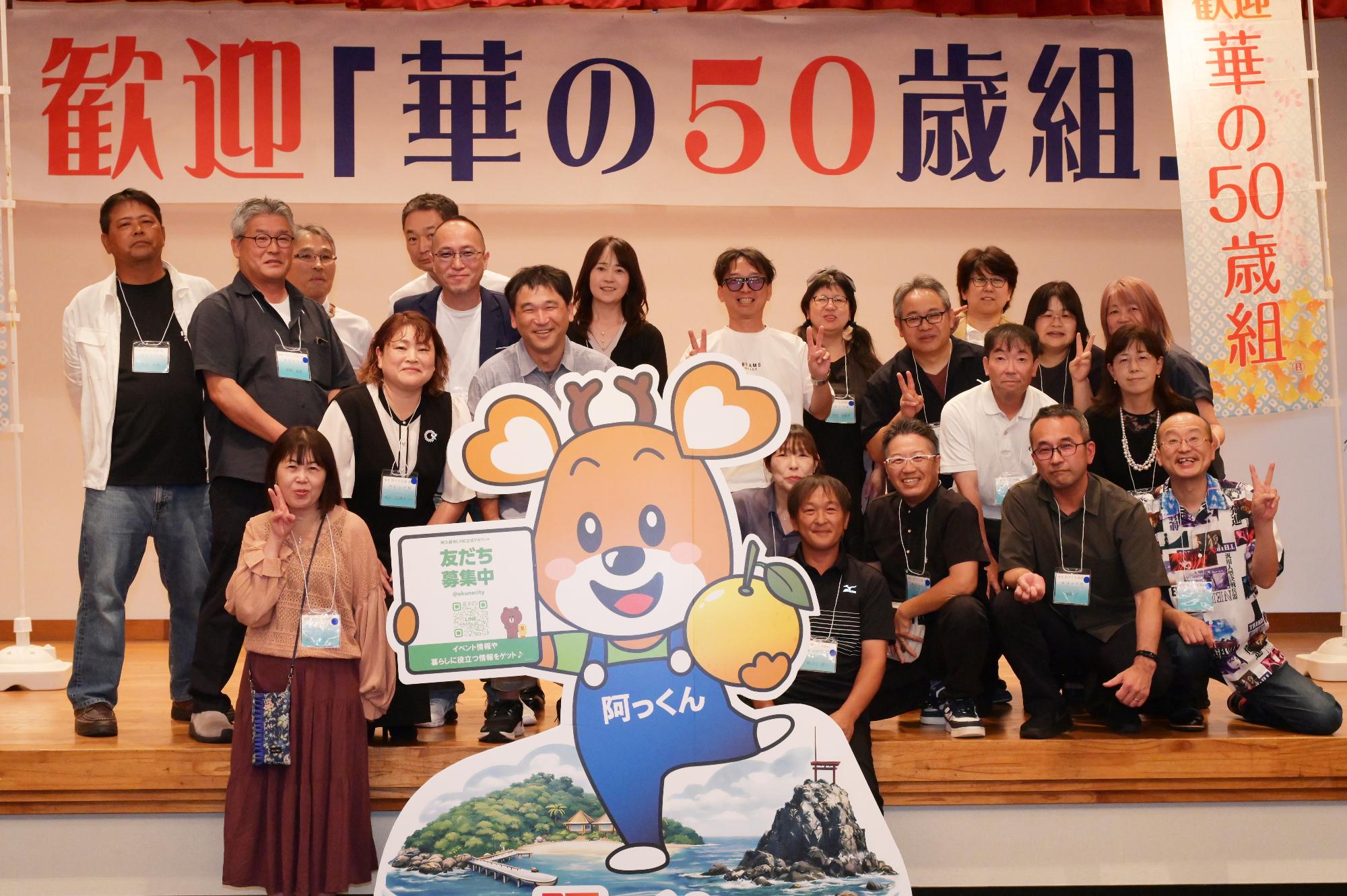 「歓迎 華の50歳組」と書かれた横断幕の前に並ぶ参加者たちの集合写真（西目小）