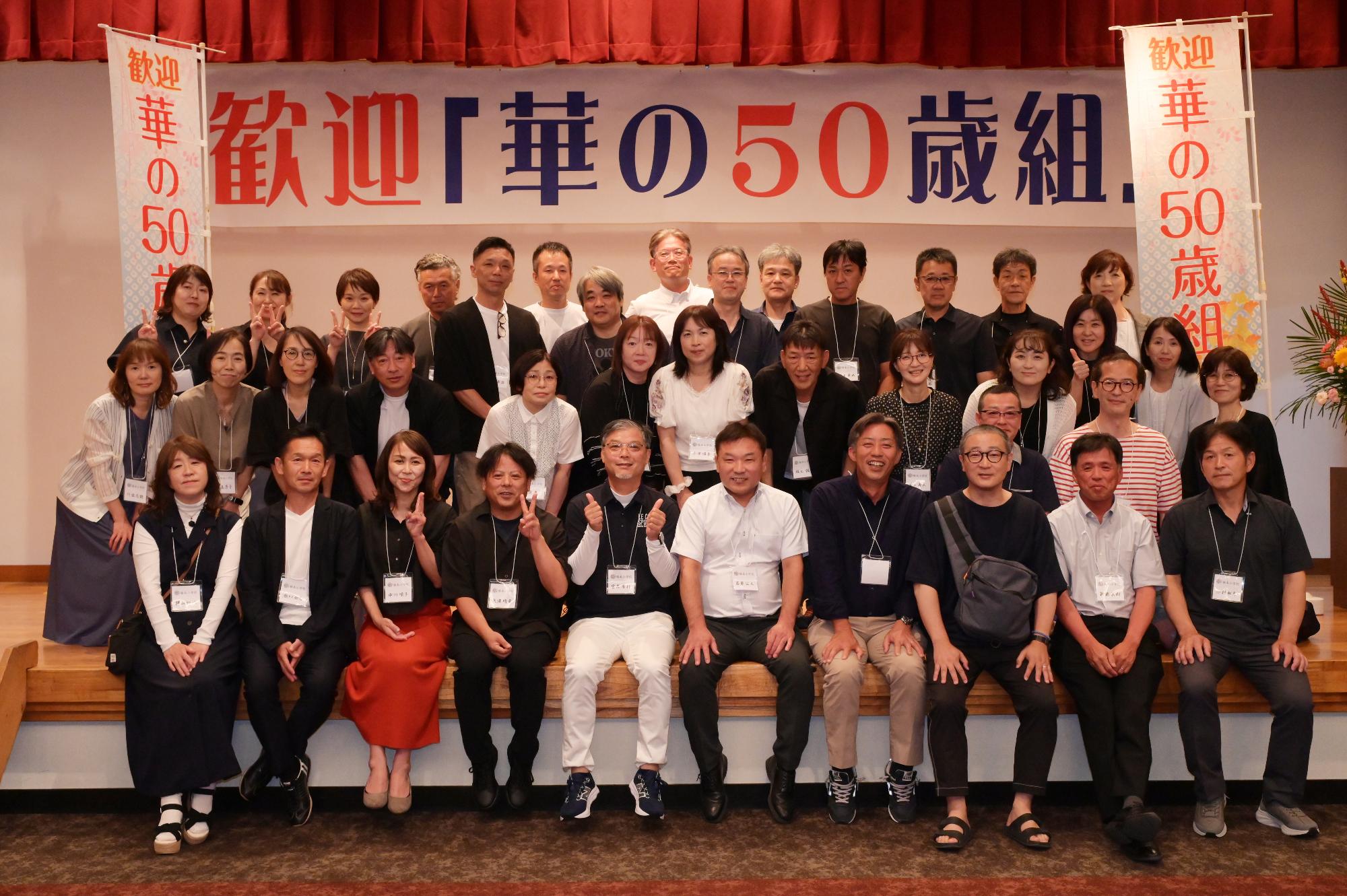 「歓迎 華の50歳組」と書かれた横断幕の前に並ぶ参加者たちの集合写真（脇本小）