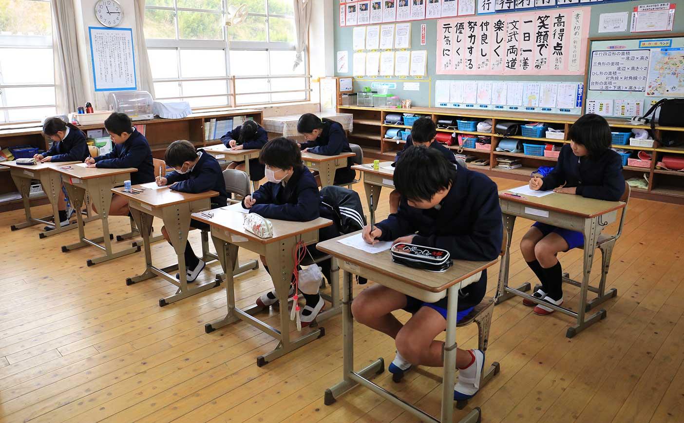 教室の机に9人の小学生が座りプリントにペンを走らせている様子の写真